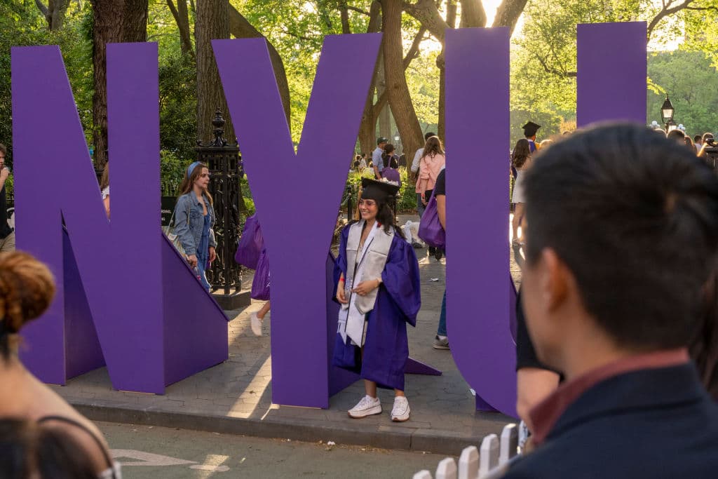 A New York University (NYU) graduate wearing a cap and gown takes photos near a large NYU sign in Washington Square Park, as seen in May 2022 in New York City. (Photo by Alexi Rosenfeld/Getty Images)