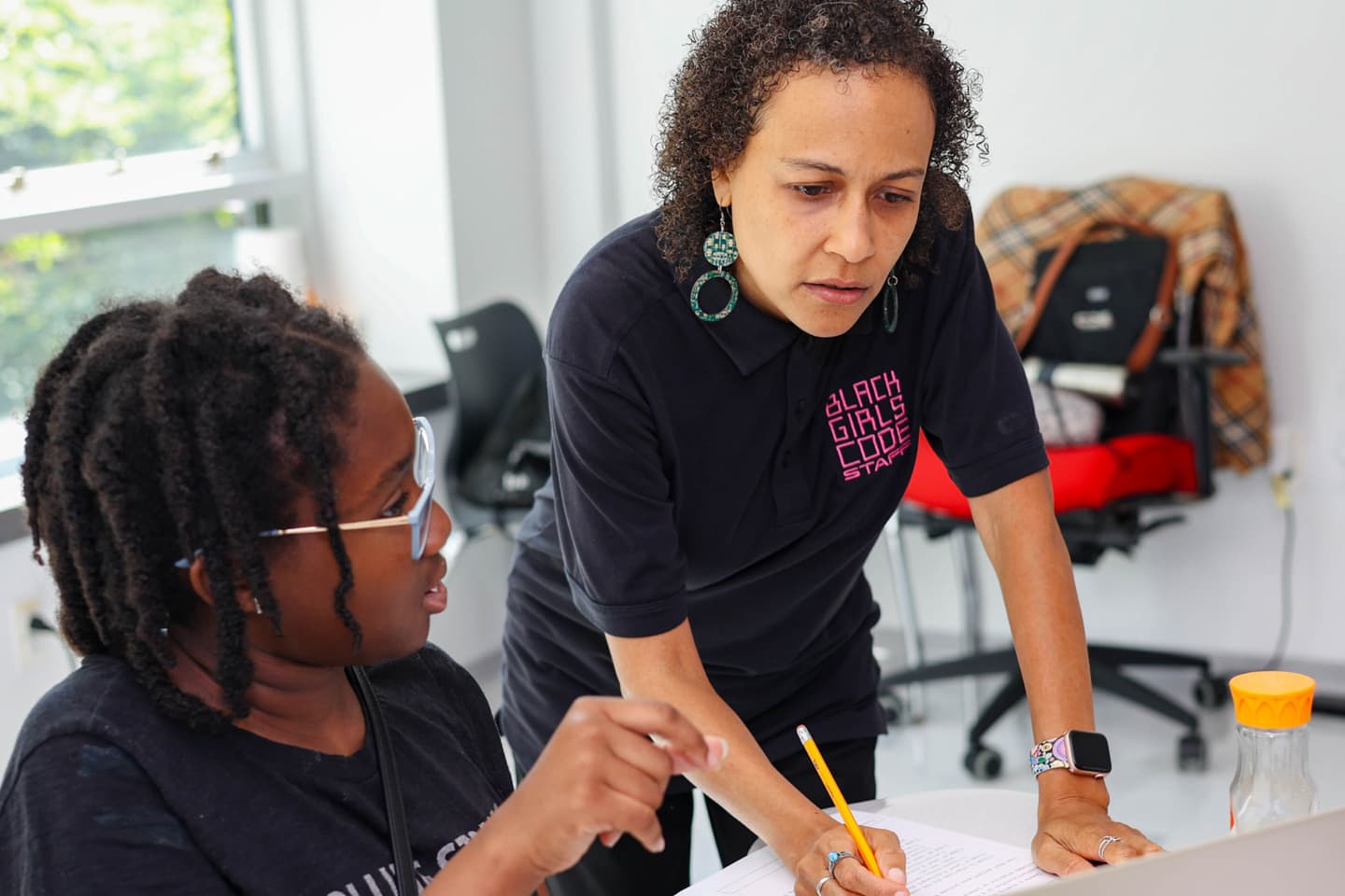 Girl with glasses is helped with an assignment by a woman with a Black Girls Code staff shirt on