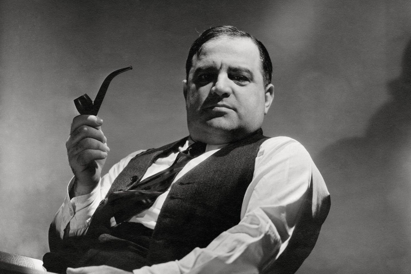 A black and white photo of a man holding a pipe, leaning back in a chair with a vest unbuttoned and a striped tie.