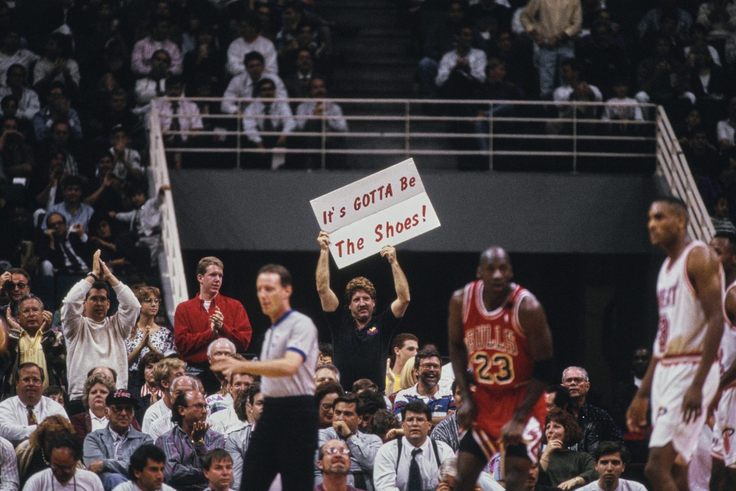 A crowd cheers near a basketball court. A fan in the background holds a sign that reads