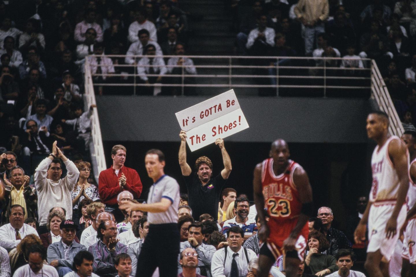 A crowd cheers near a basketball court. A fan in the background holds a sign that reads 
