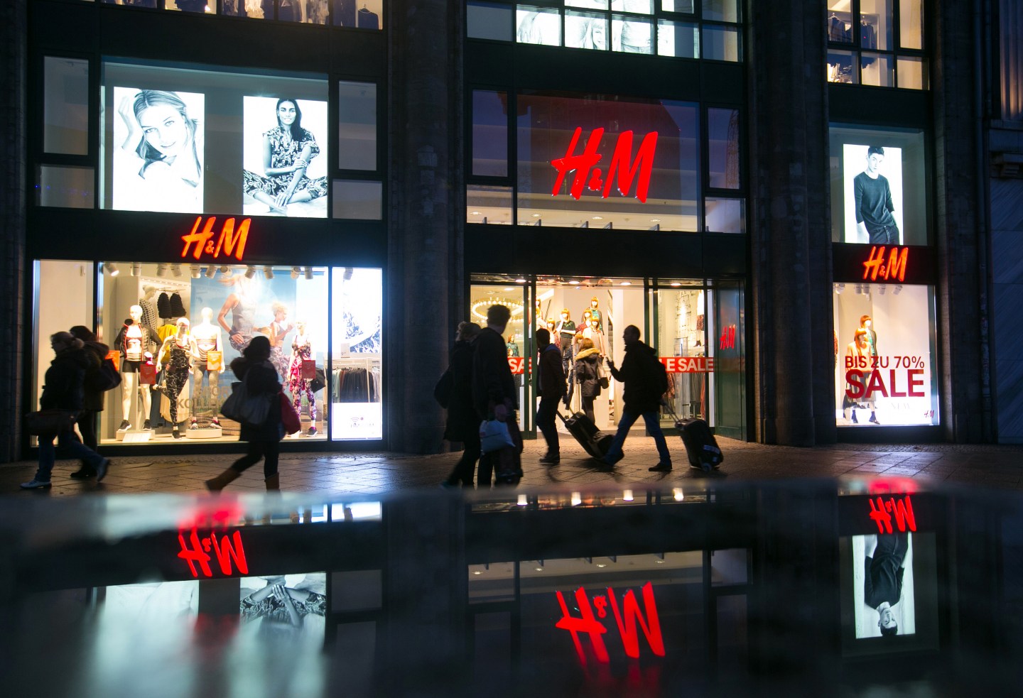 Pedestrians pass an illuminated Hennes &amp; Mauritz AB (H&amp;M) clothing fashion store in Berlin, Germany, on Wednesday, Jan. 7, 2014. As European Central Bank policy makers gather for their first meeting of 2015 today, the backdrop is a 0.2 percent annual drop in consumer prices, the first in more than five years. Photographer: Krisztian Bocsi/Bloomberg