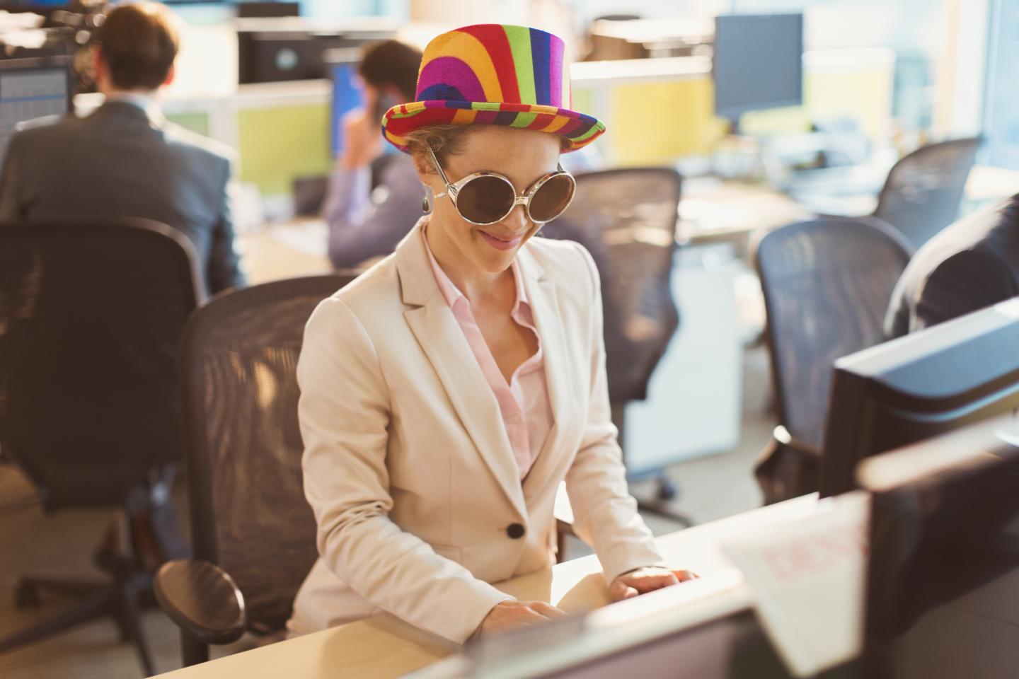 Playful businesswoman wearing silly sunglasses and striped hat at computer in office