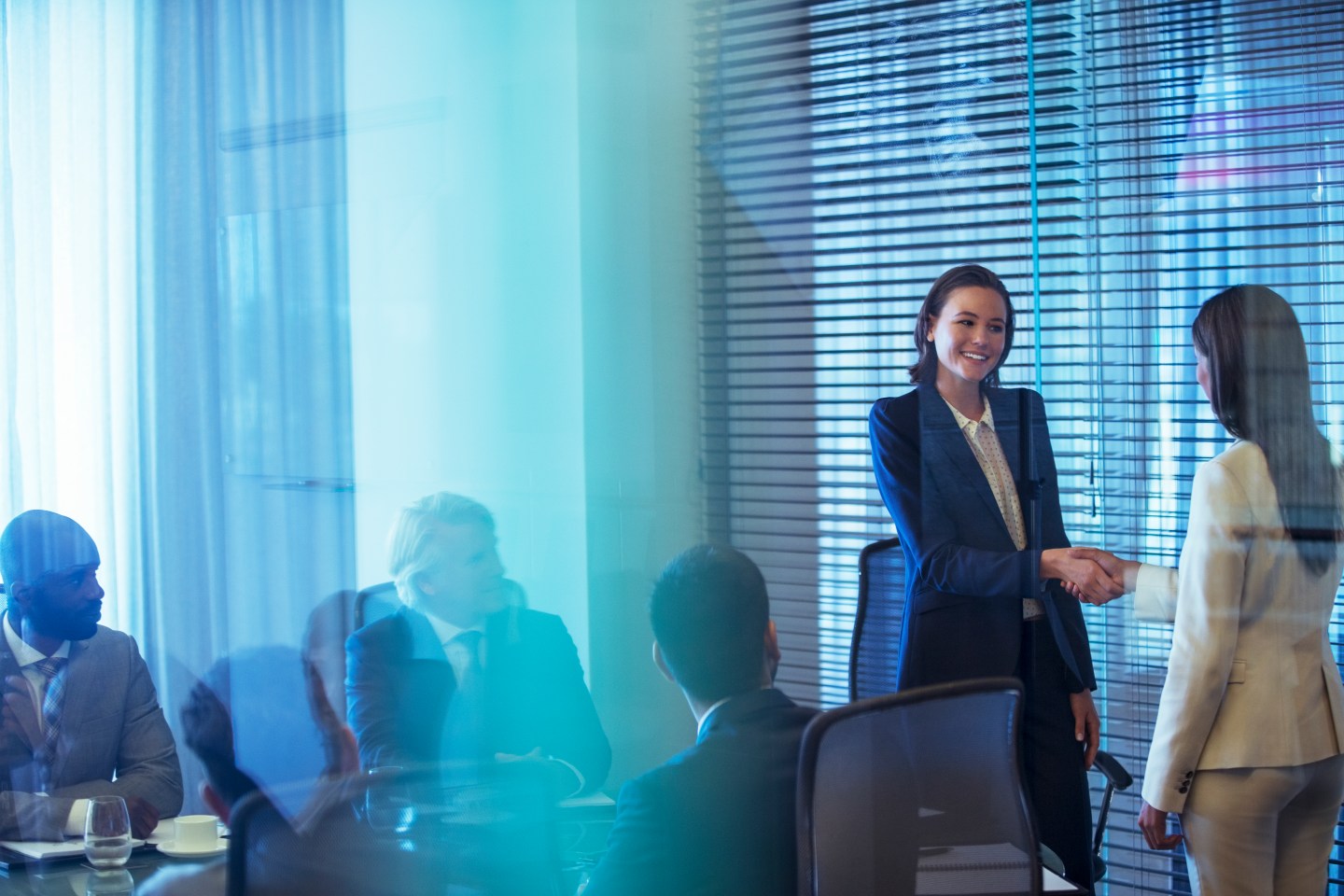 Two businesswomen shaking hands in conference room, colleagues watching