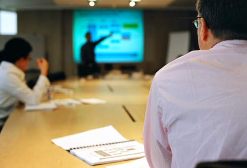 A scene in a meeting where a businessman presents with digital projection on a screen.