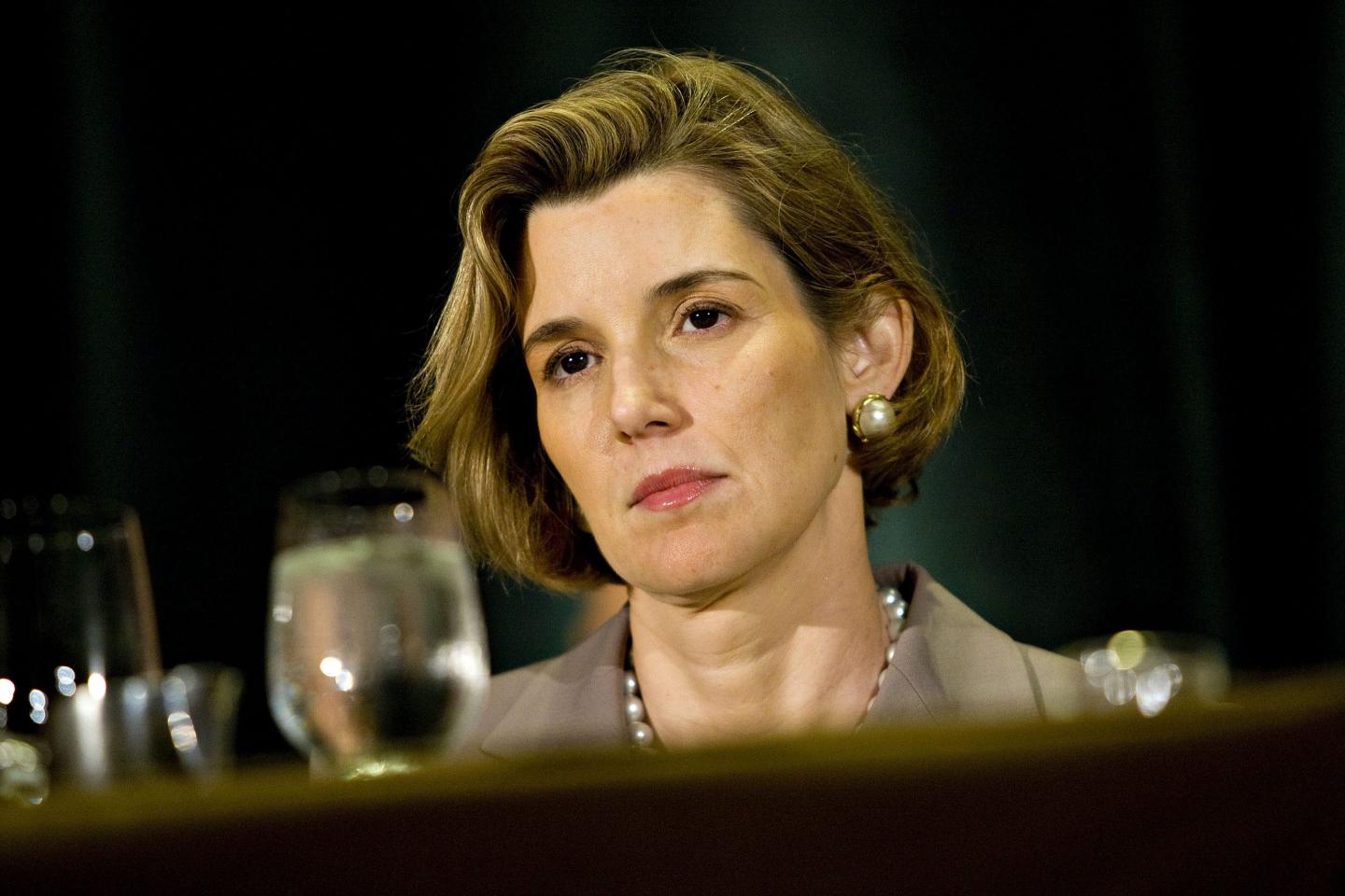 A white woman with a beige blazer sits behind a table with a glass of water on it