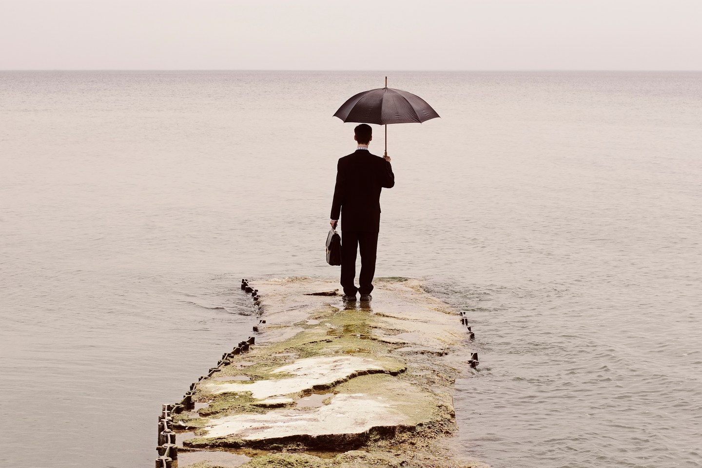 Man in suit holding umbrella and briefcase, standing at end of sinking pier