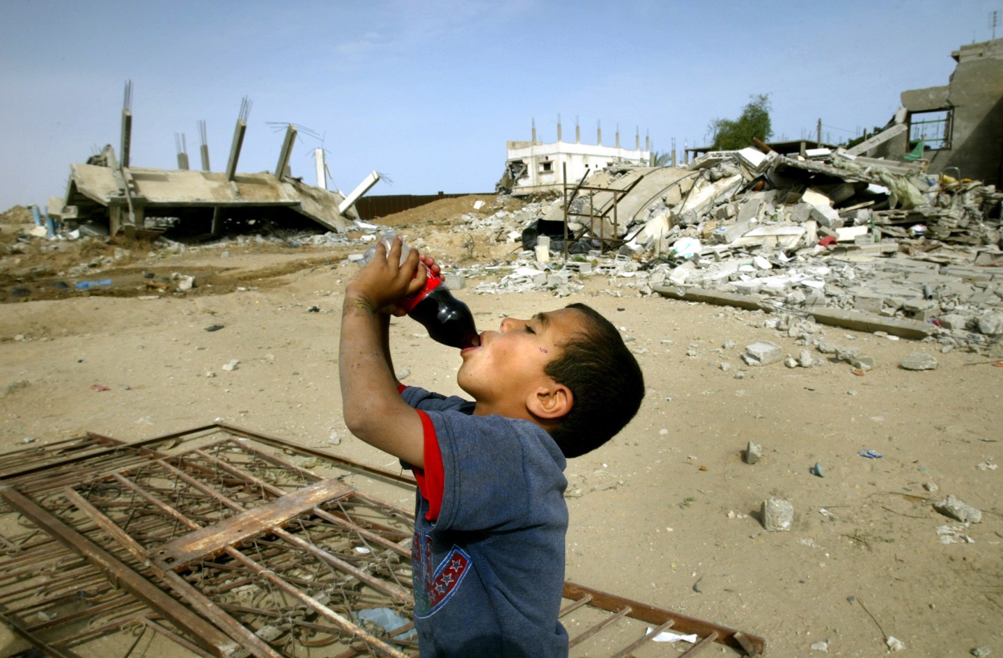 PALESTINIAN BOY DRINKS COCA-COLA NEXT TO HOUSES DESTROYED AT RAFAH REFUGEE CAMP.
