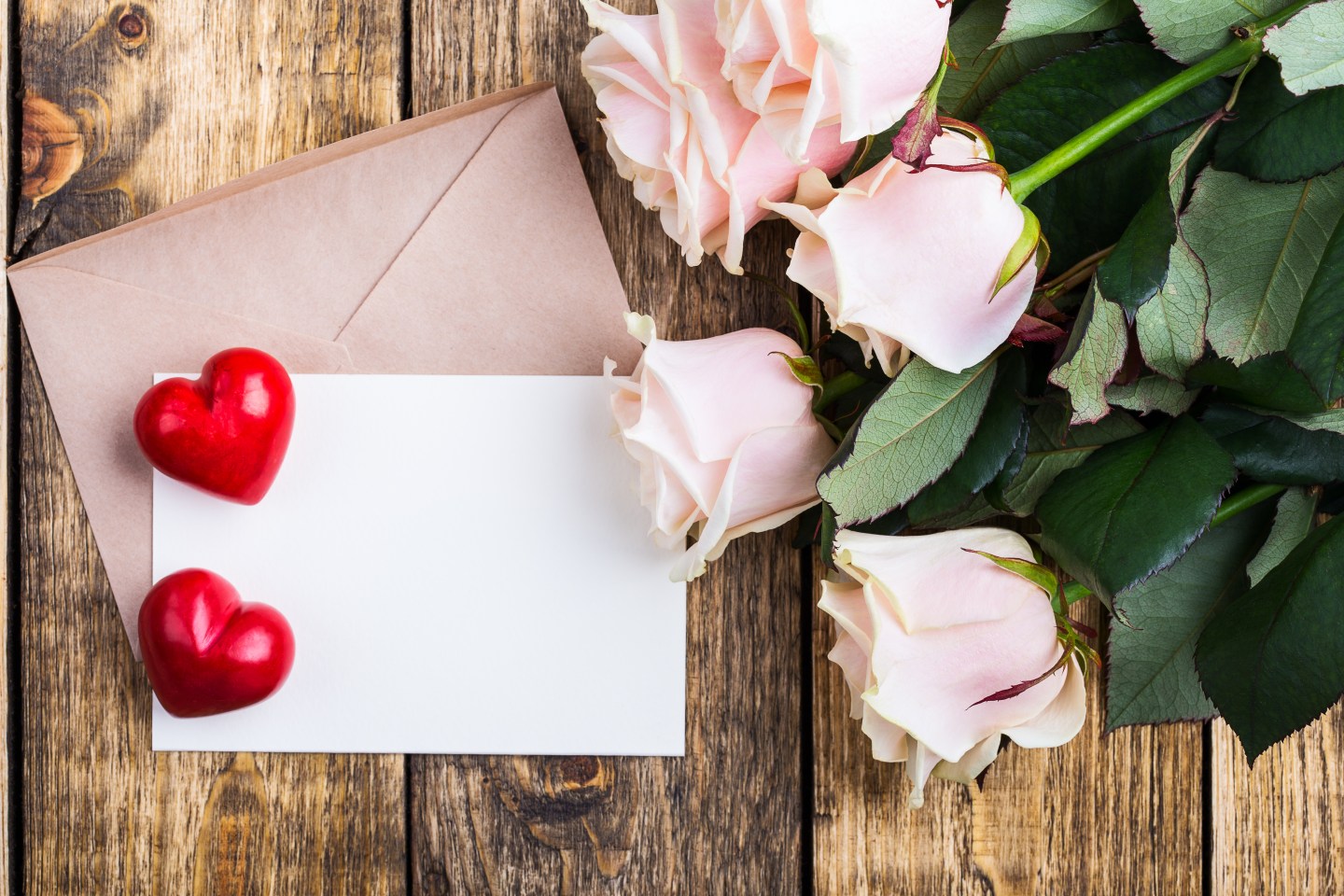 Pink roses and two red hearts on a rustic wooden board. Envelope and empty blank card for message on Valentine's Day