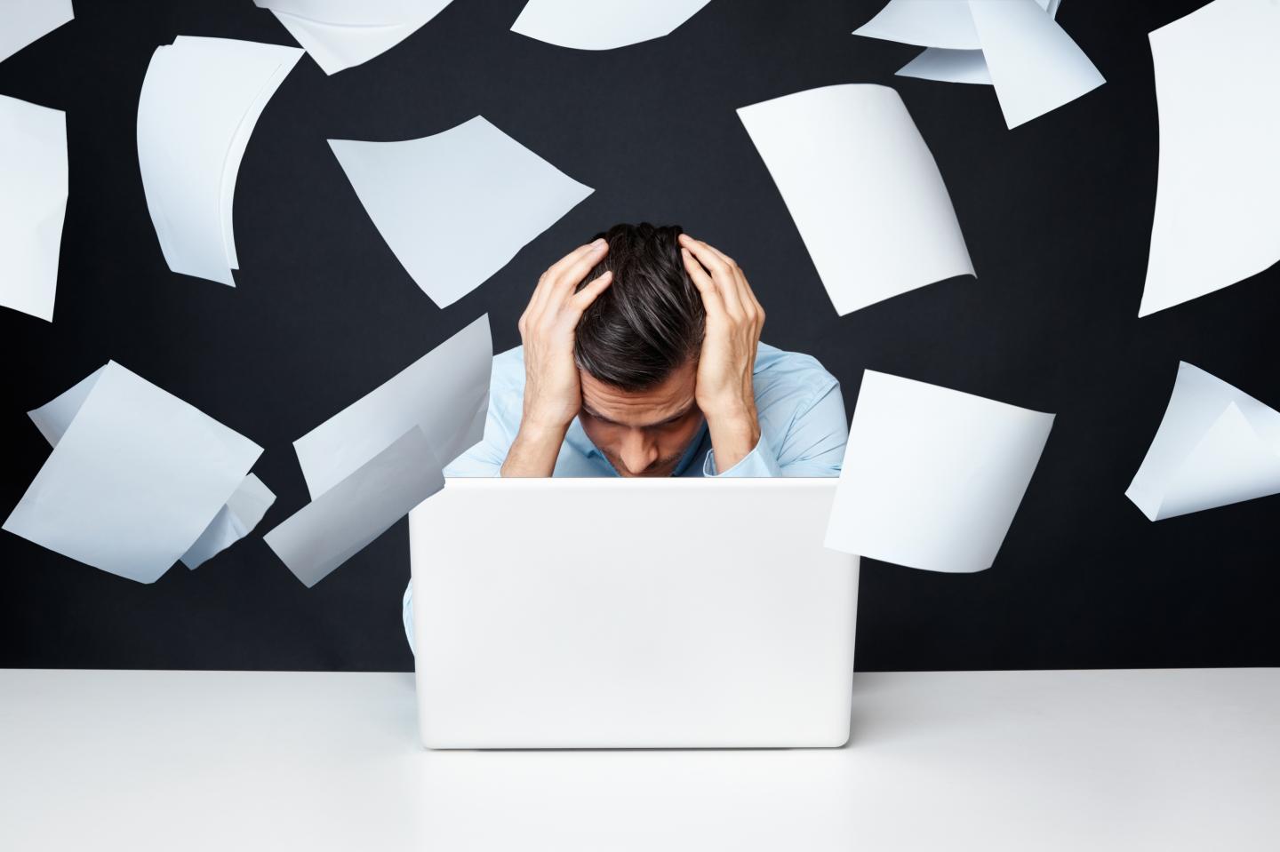 Man with hand on head looking at laptop with papers flying