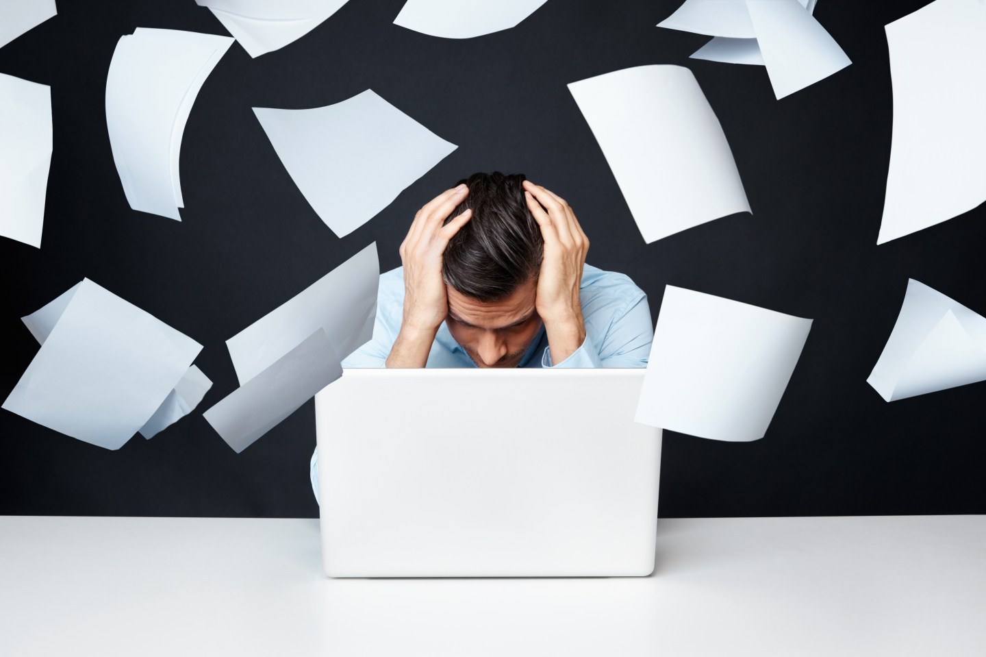 Man with hand on head looking at laptop with papers flying