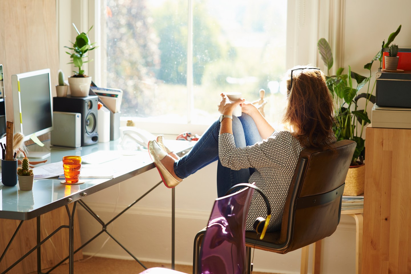 Pensive woman looking through window with feet up on desk in sunny home office