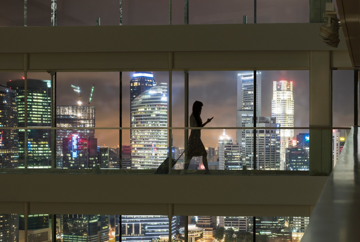 Young woman using smartphone and pulling suitcase, city skyline in view