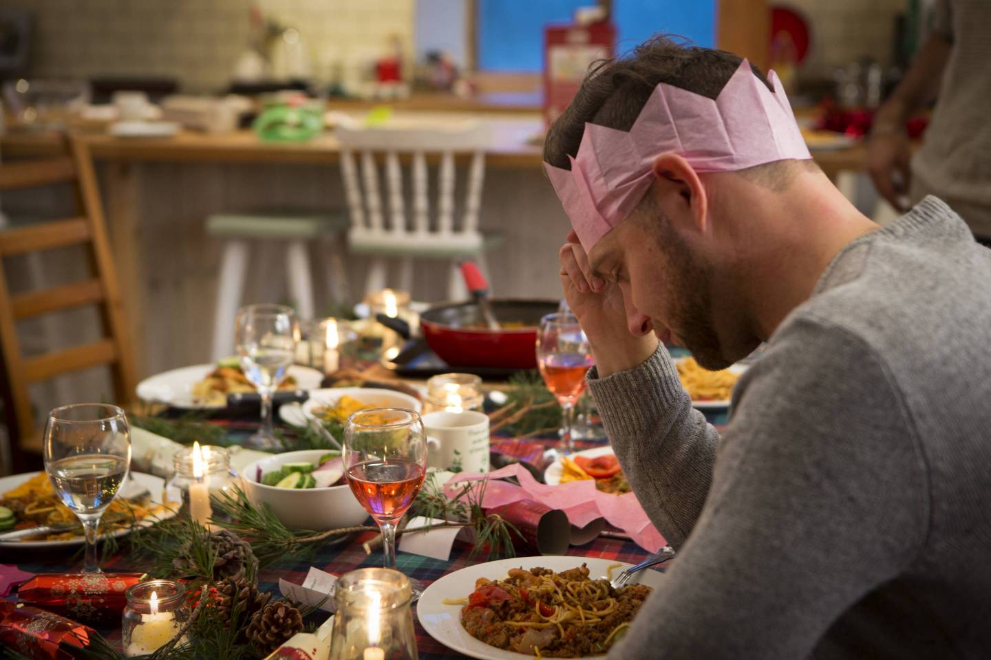 A man sits at a dinner table at Christmas looking sad as his head is lowered and resting on his hand. He sits with a pink hat on, alone with a plate of food in front of him.