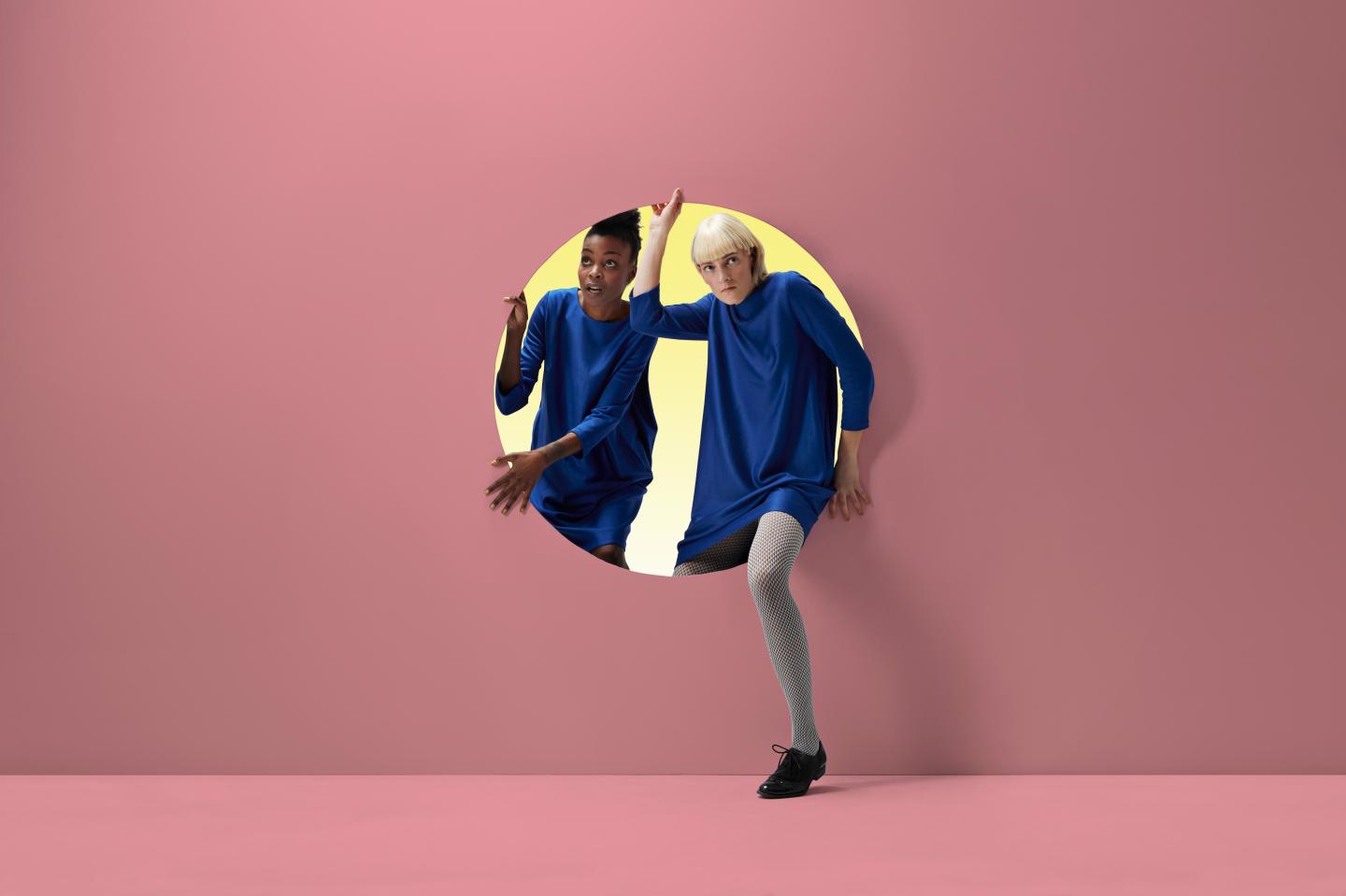 Two women peeking out of round opening in coloured wall