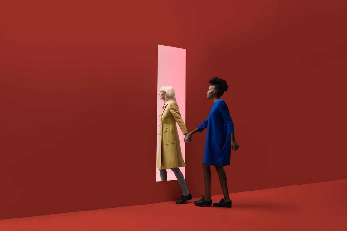 Two women holding hands, walking threw rectangular opening in coloured wall