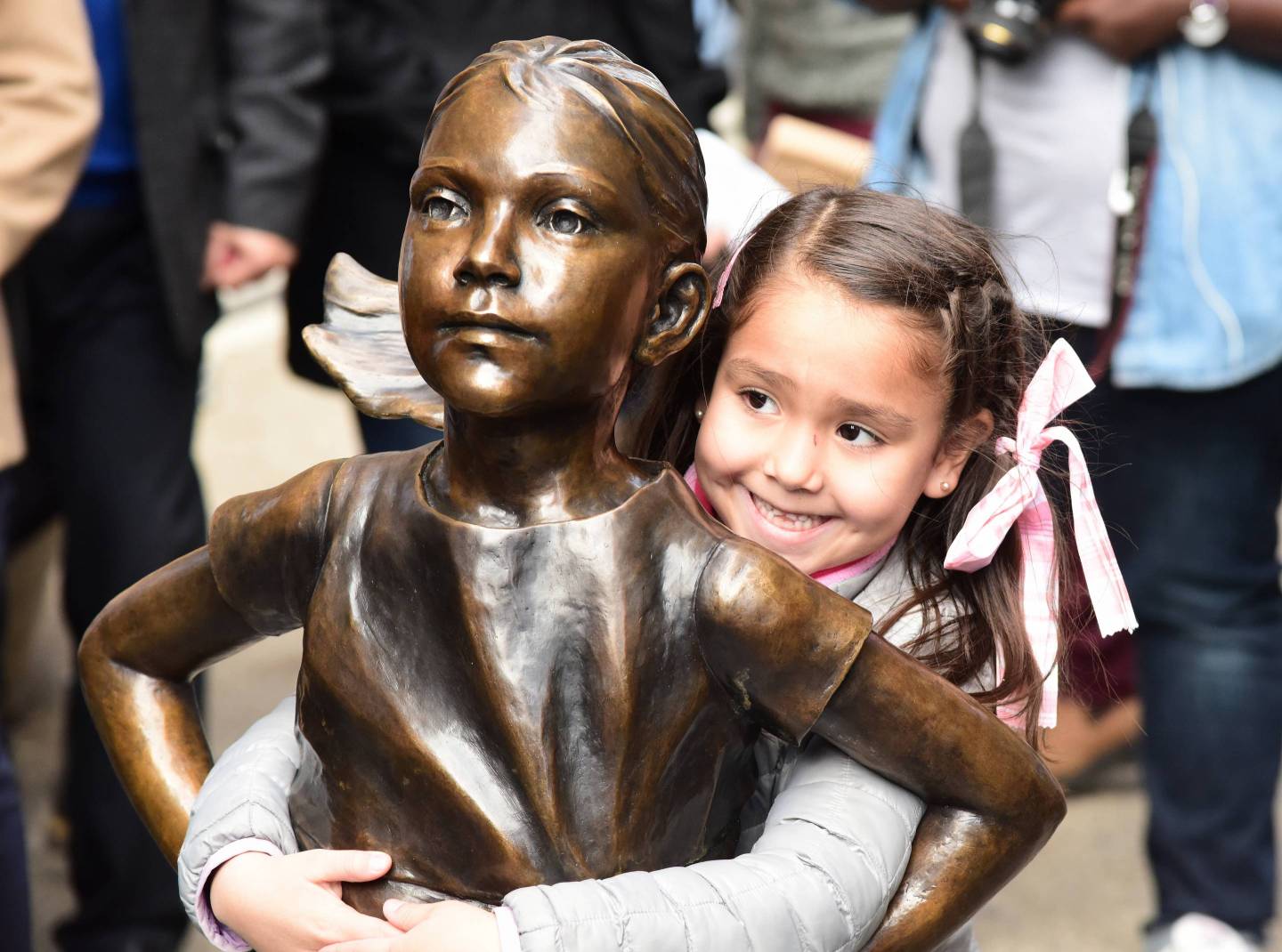 Young girl in pigtails hugs the waist of the Fearless Girl statue.