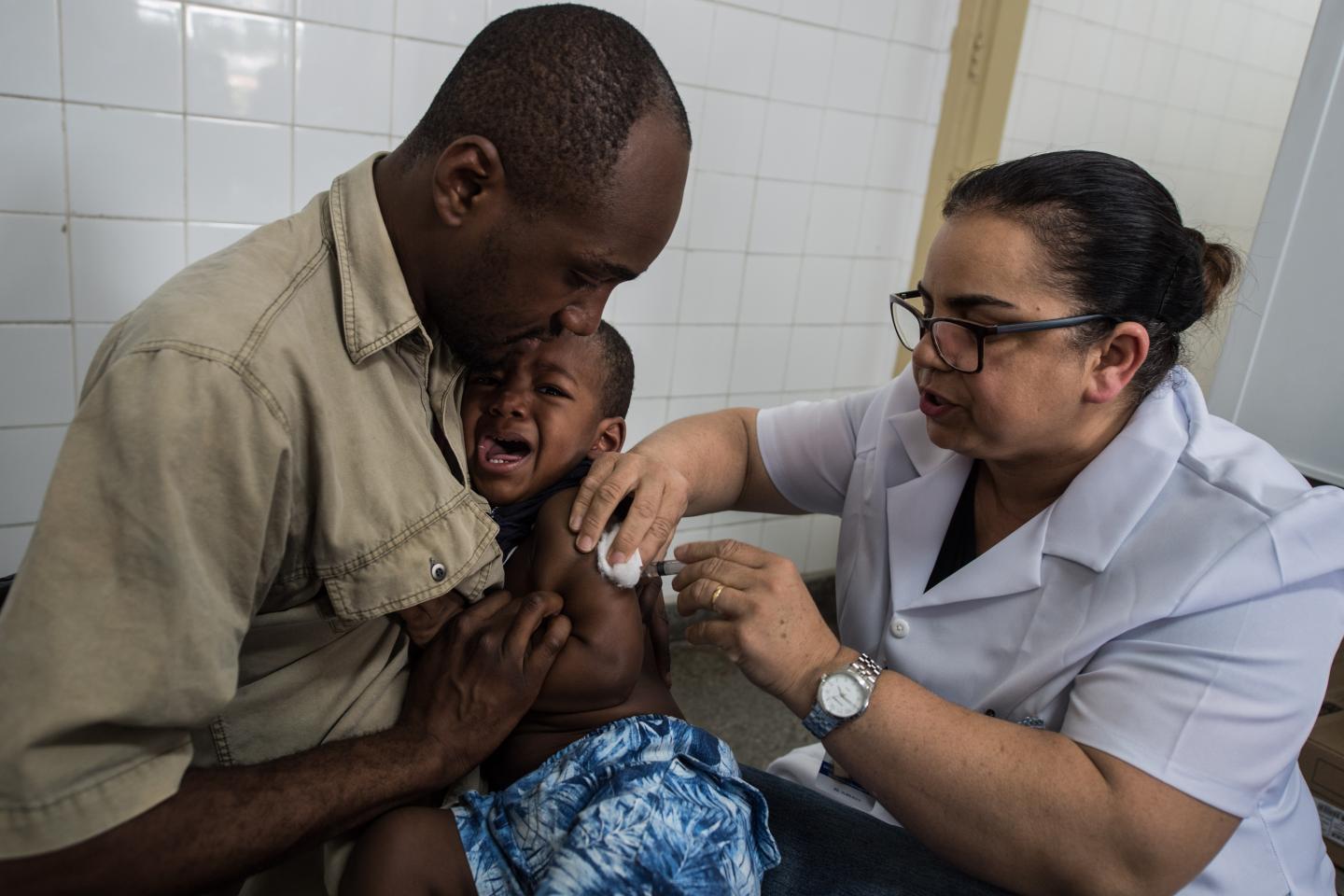 A man holds a boy who is crying as he is vaccinated against yellow fever by a nurse.