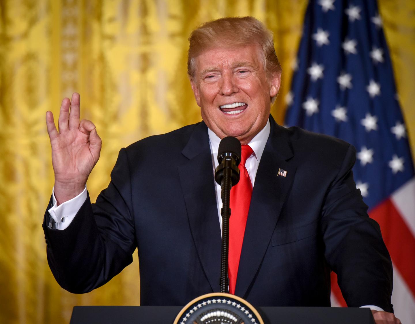 President Donald Trump stands in front of an American flag with his hand raised and his mouth open at a podium in the White House.