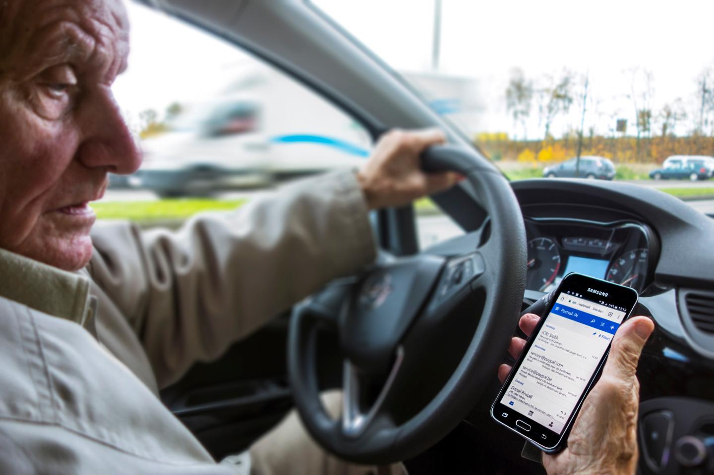 Elderly man at steering wheel checking messages on smart phone while driving car on road.