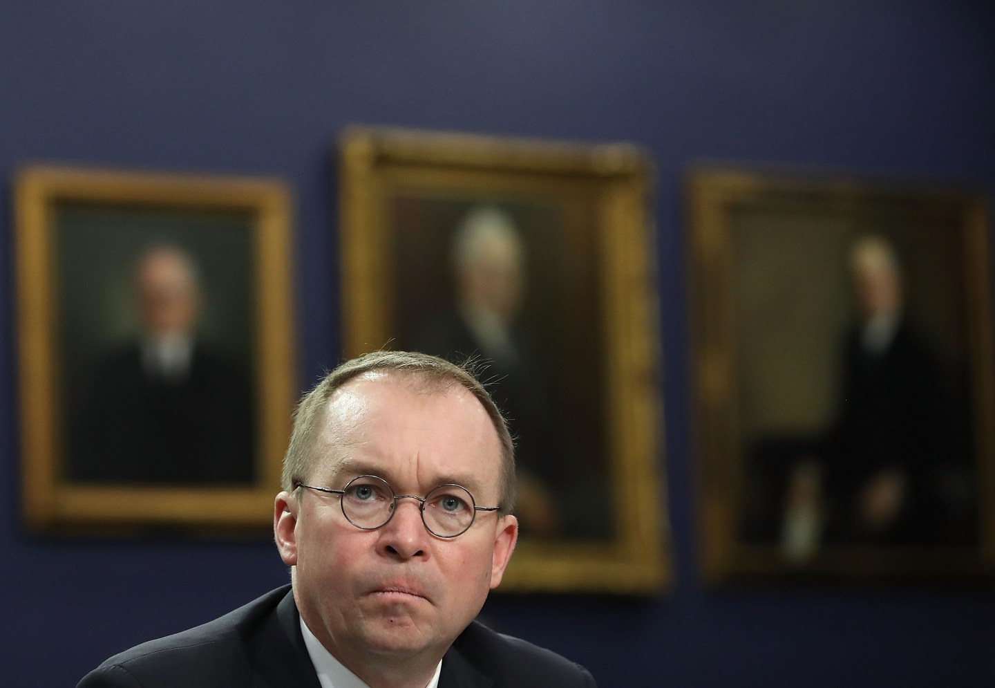 CFPB and Office of Management and Budget director Mick Mulvaney stares at the camera with a frown on his face. he wears glasses and sits in front of three large portraits which are out of focus.