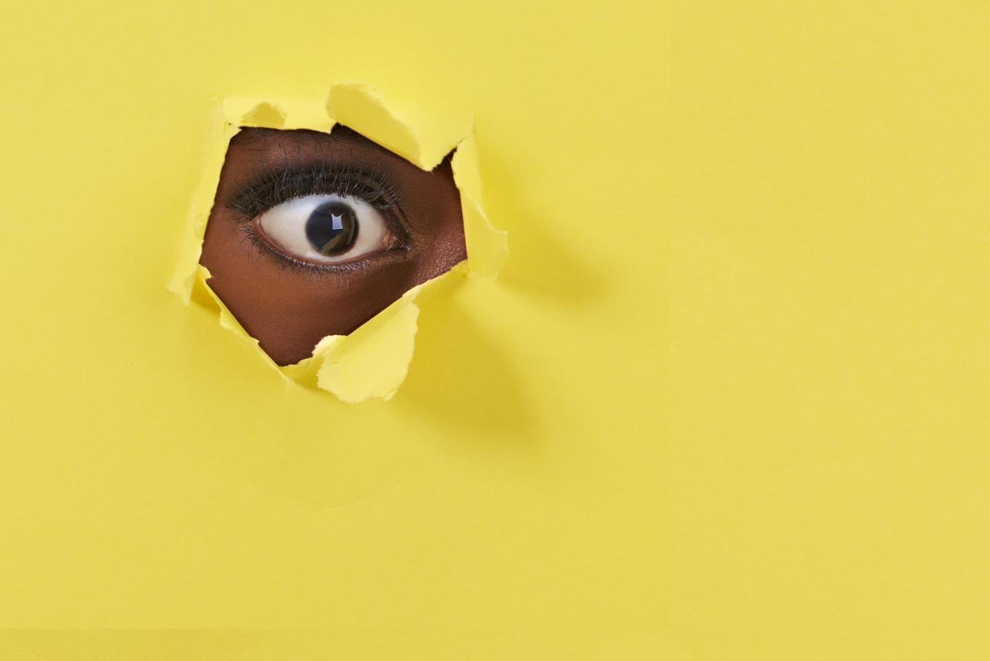 A view of a woman's eye looking through a hole in yellow paper