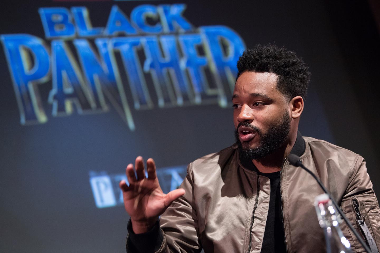 Ryan Coogler speaks to someone out of the frame, his hand raised as he illustrates his point in front of a large projection of the 'Black Panther' movie logo.