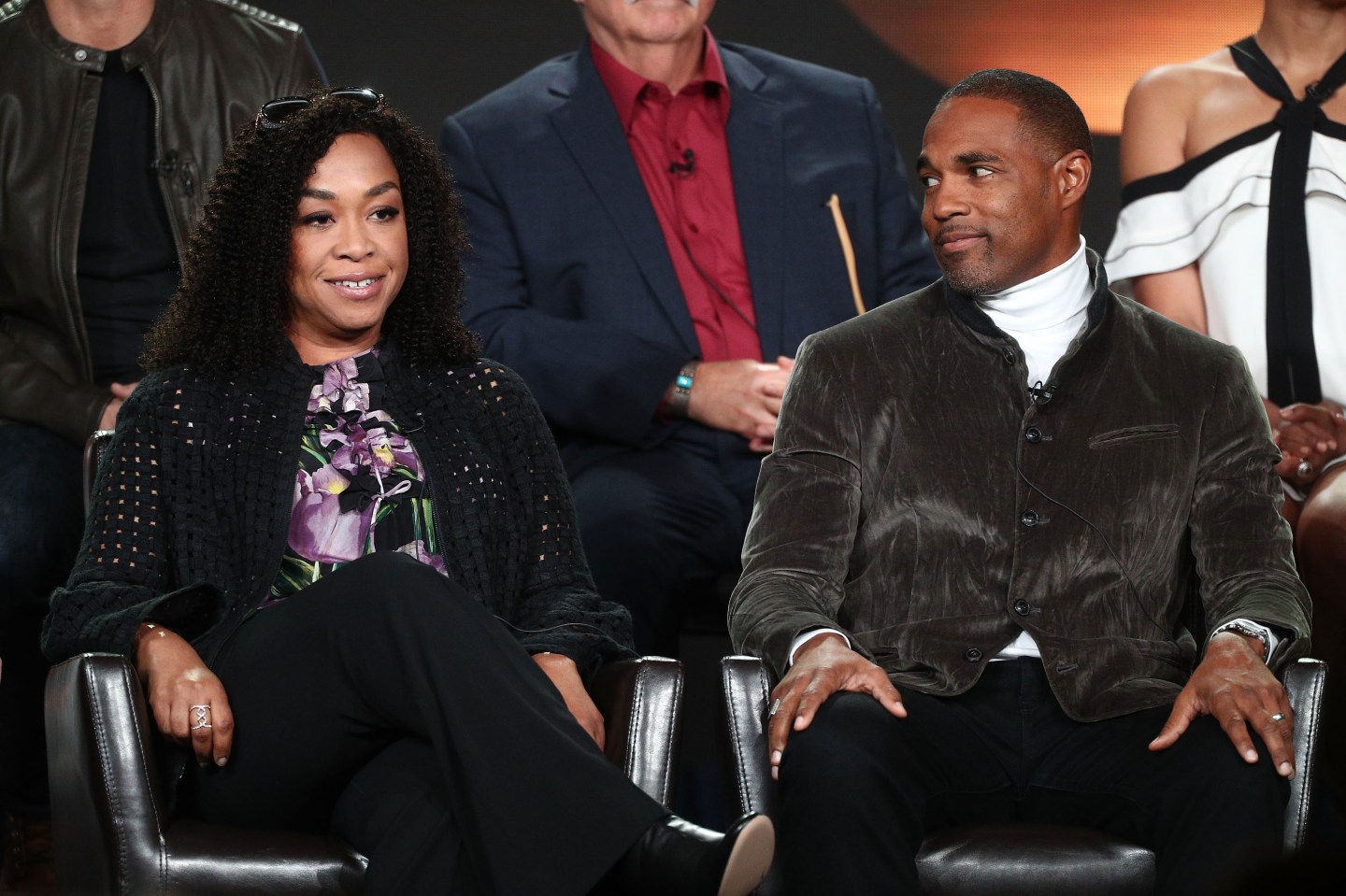 Shonda Rhimes sits in a chair speaking on stage, actor Jason George sits on her left and looks enthralled by what she is saying.