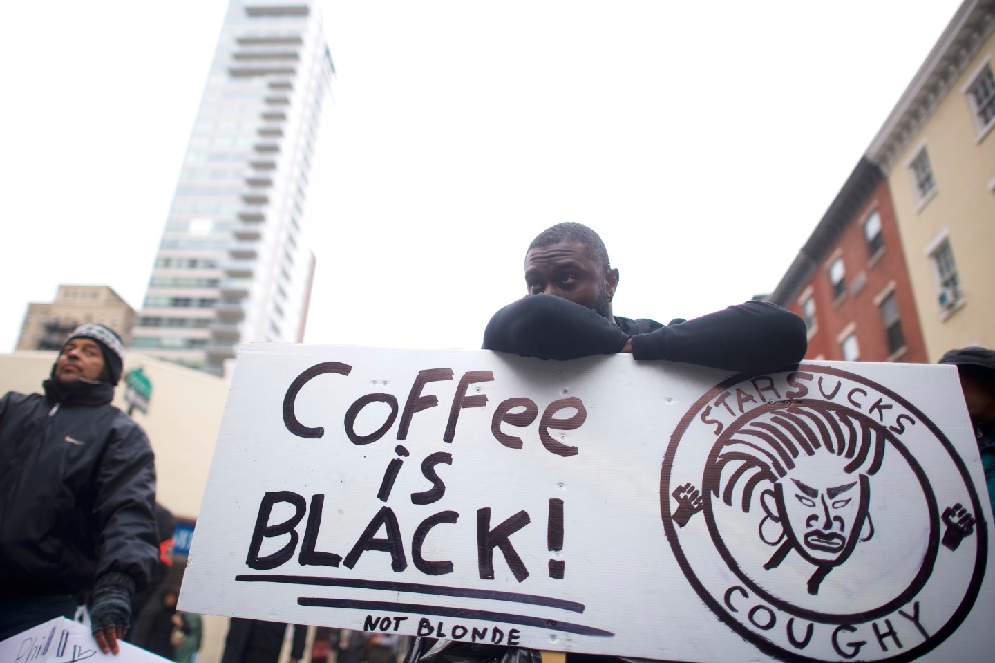 A man rests his head on top of his arms which are supported by a large picket sign that says "Coffee is Black!" during a protest in Philadelphia.