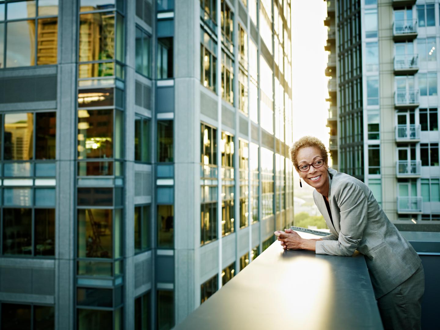 Businesswoman leans on the edge of deck smiling, the sun reflects through two tall glass buildings behind her