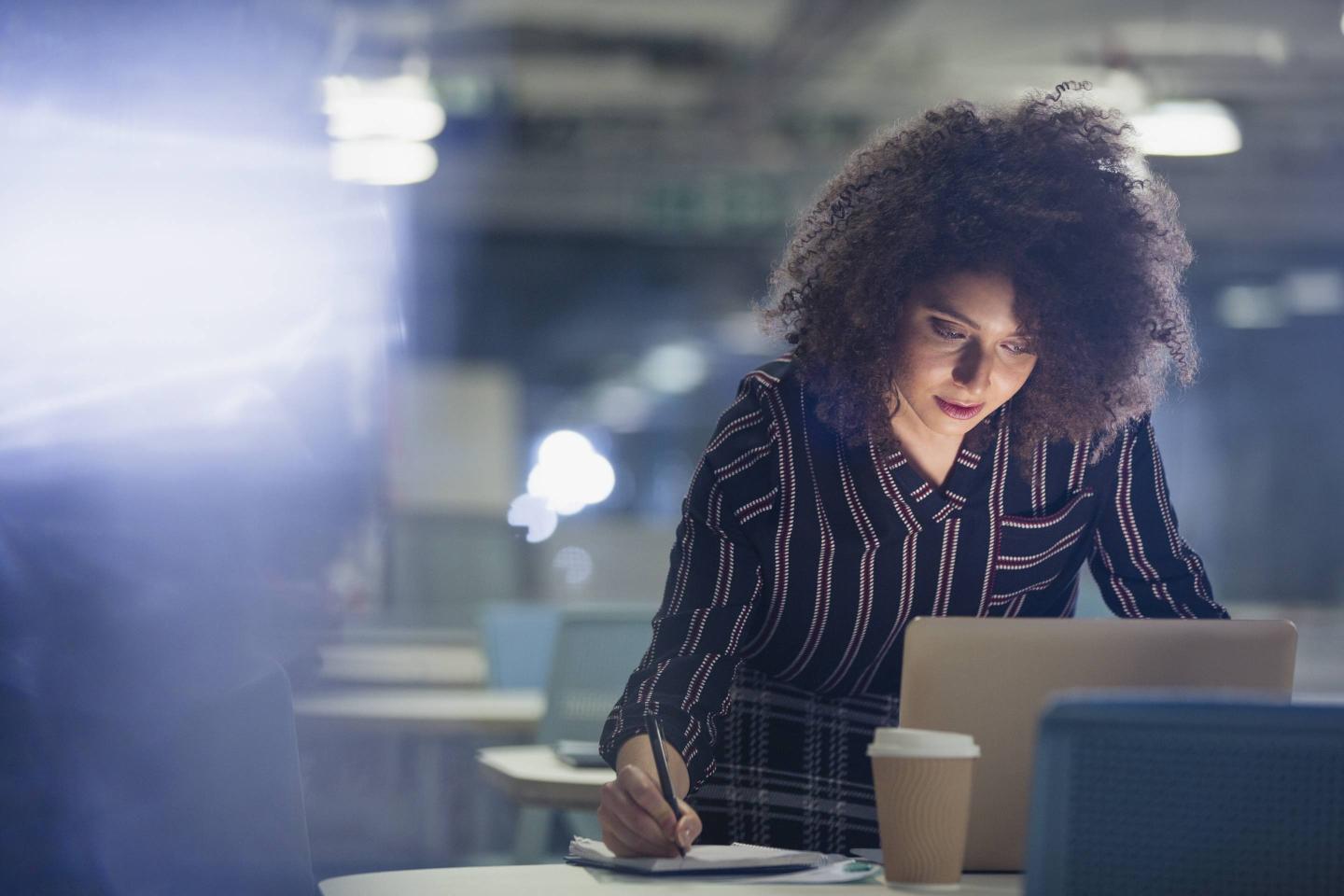 Focused businesswoman working late at laptop, taking notes in dark office