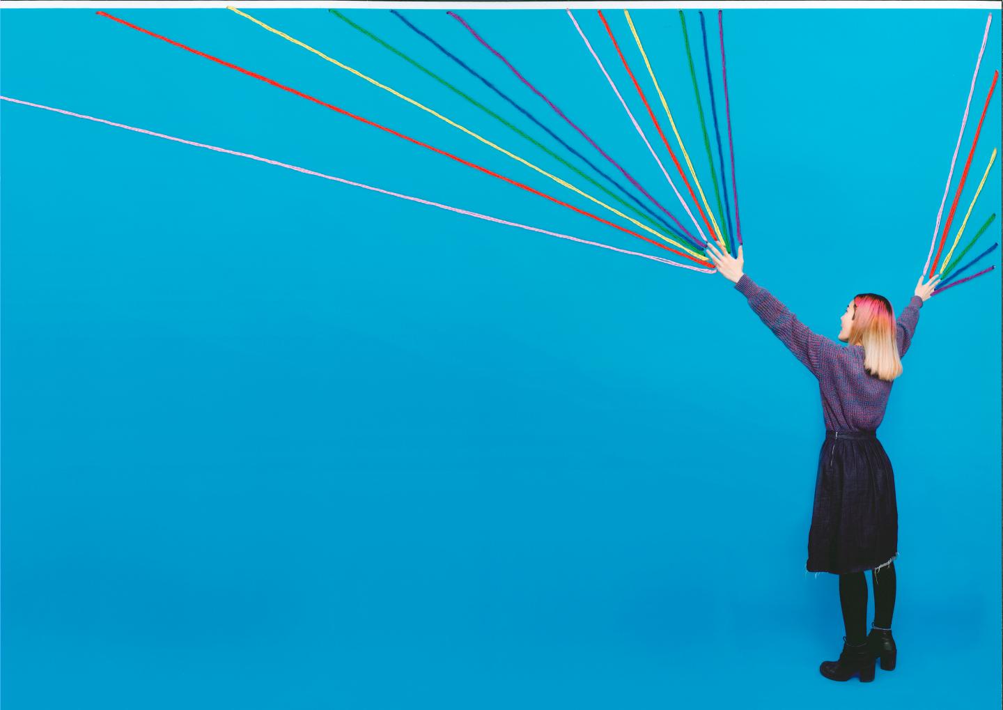 A woman faces away from the camera. She has her arms raised and rainbow strands shoot out of her fingertips.