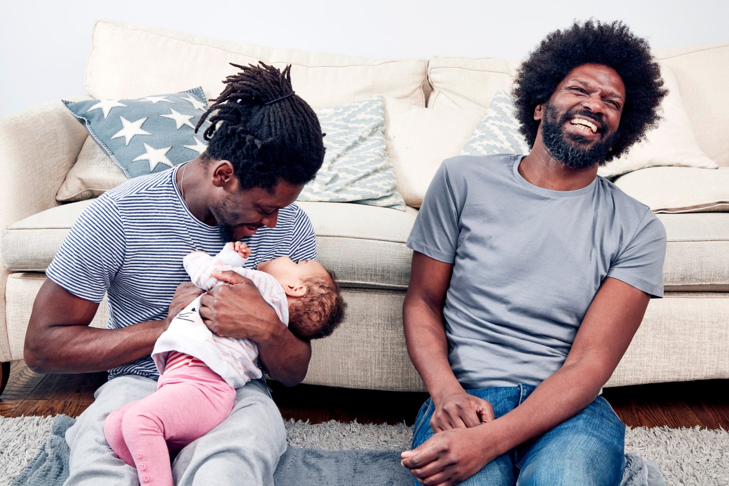Two black fathers are beaming as they sit on the floor and play with their daughter