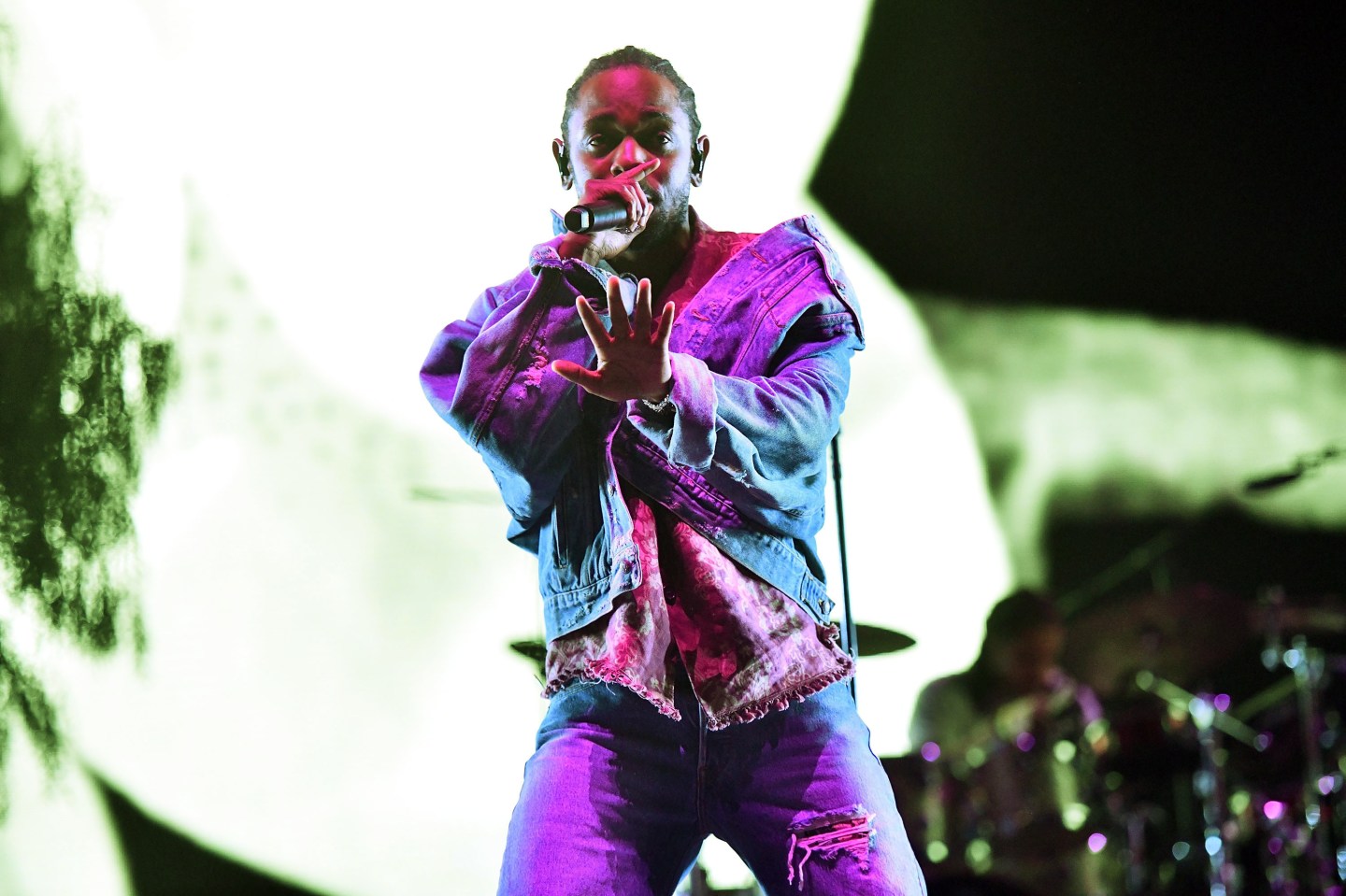 Kendrick Lamar raises the microphone to his mouth with one hand and raises his other hand in a "stop" motion.