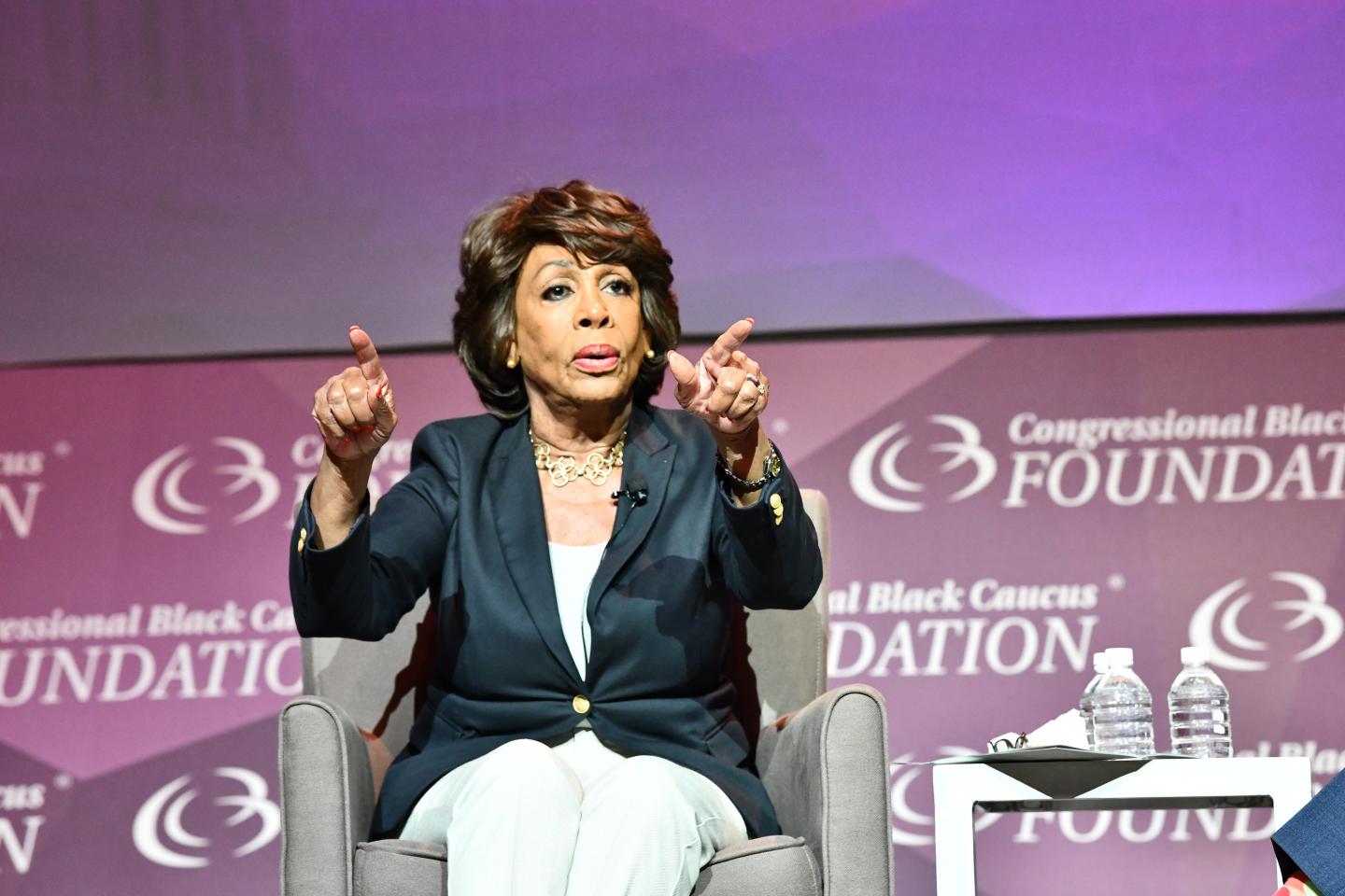 Maxine Waters sits on stage in front of a purple background and points out at the audience with both hands.