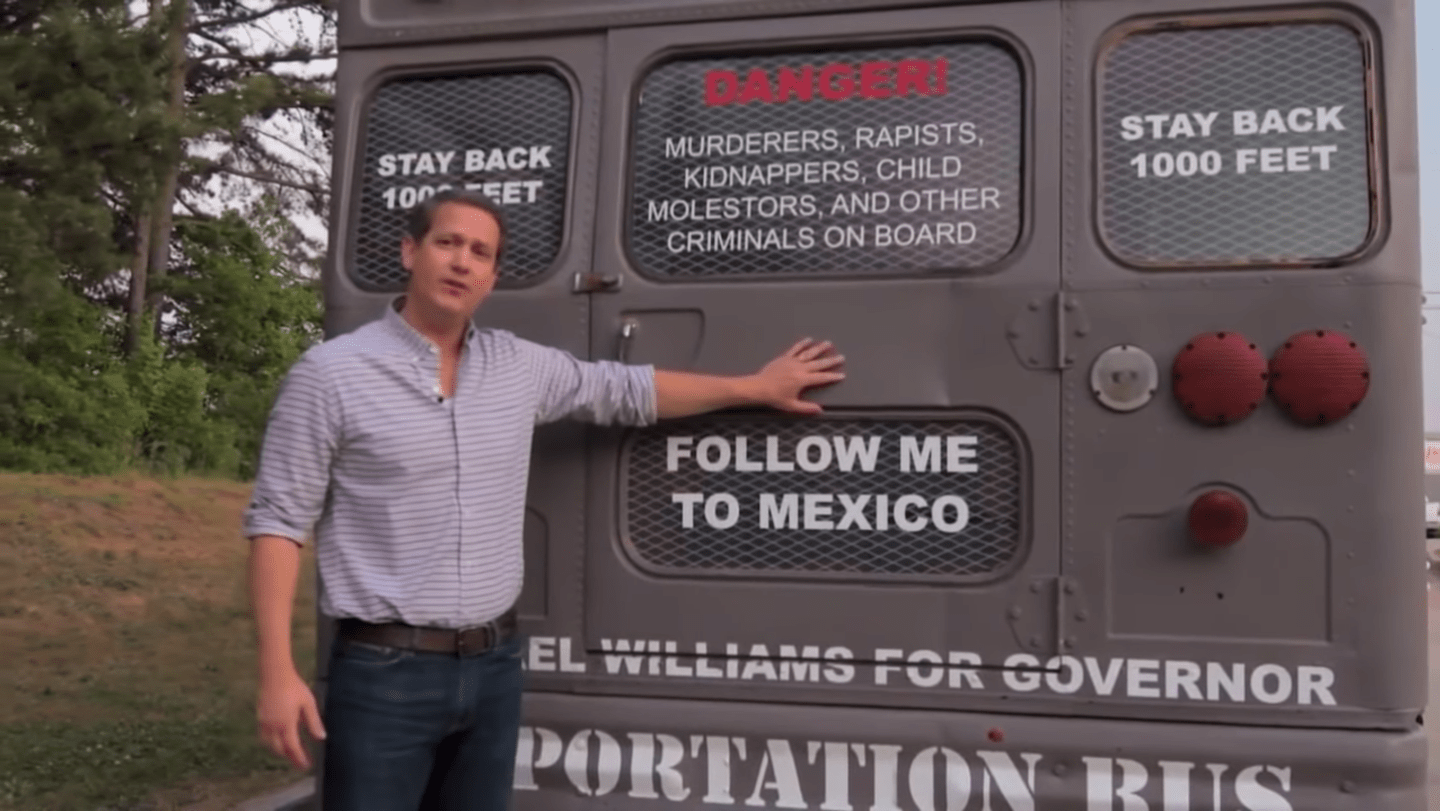 Georgia state Sen. Michael Williams stands in front of a gray bus and places his hand on the back door over the words "FOLLOW ME TO MEXICO"