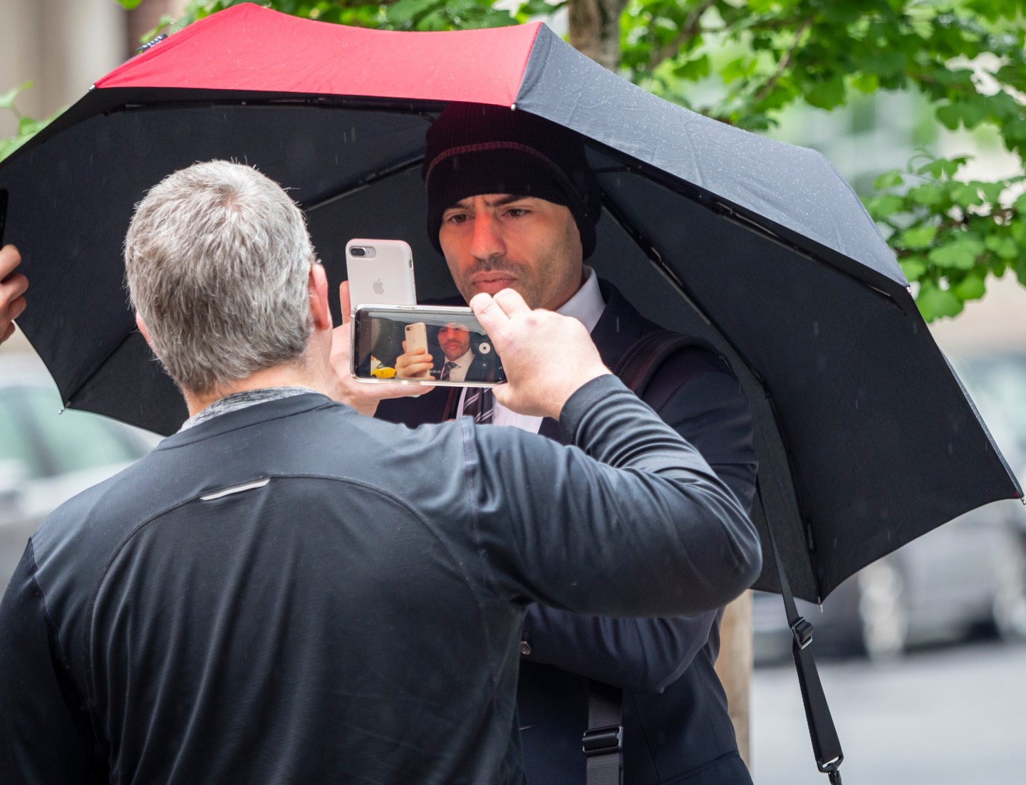 A man in a black ski cap with a red and black umbrella takes a video of another man who is recording a video of the man in the hat on his phone.
