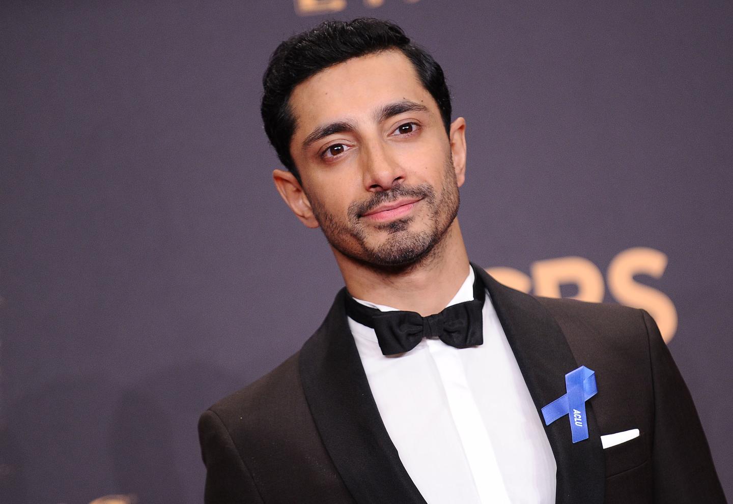 Riz Ahmed poses at the Emmy Awards. he wears a bowtie and a blue ribbon.