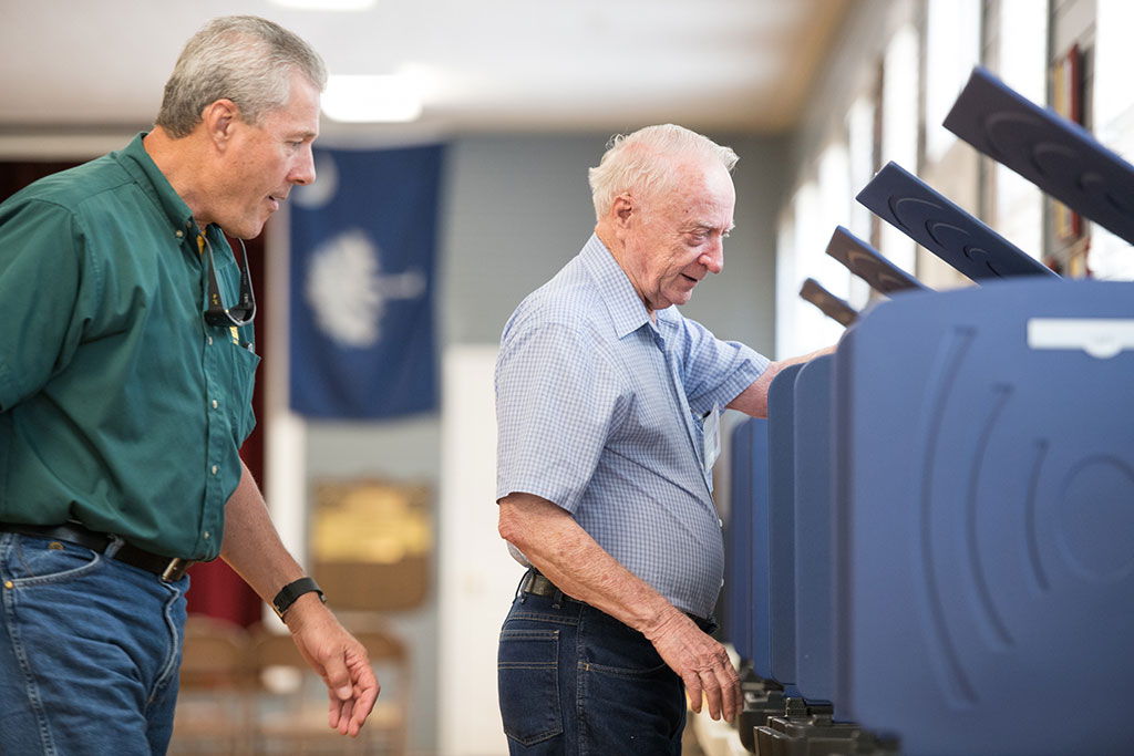 An older man stands at a voting booth while another man watches.