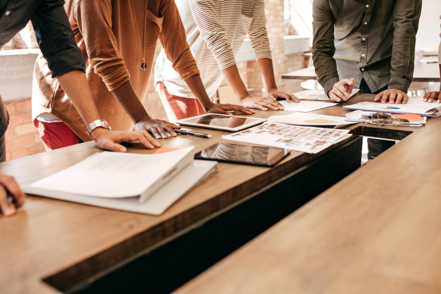 four people stand around a table, leaning both arms on it above stacks of papers.