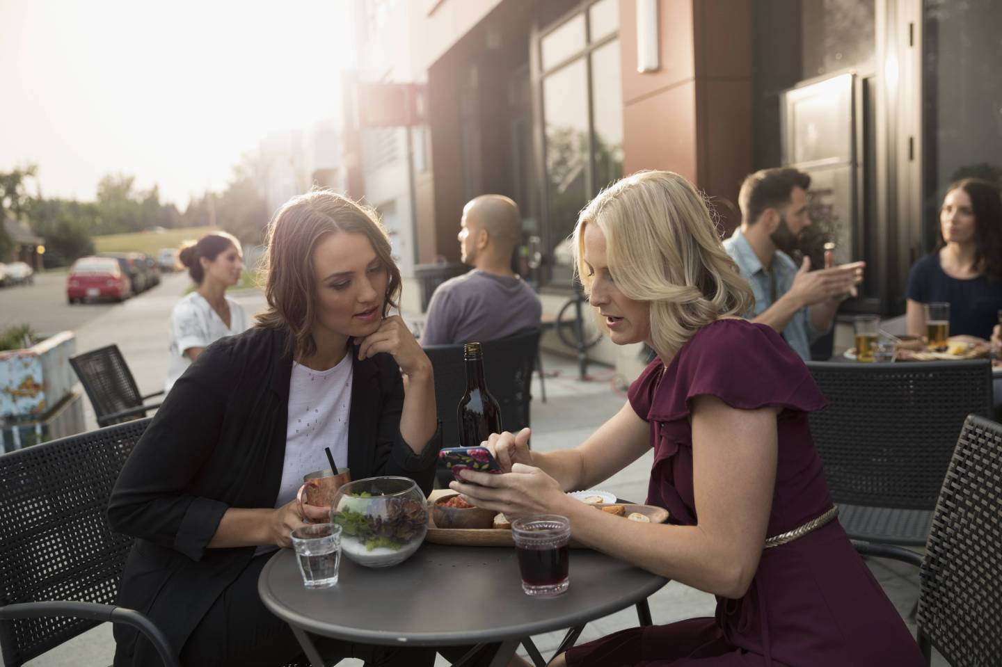 Women friends enjoying wine and cocktail, texting with cell phone at sidewalk cafe