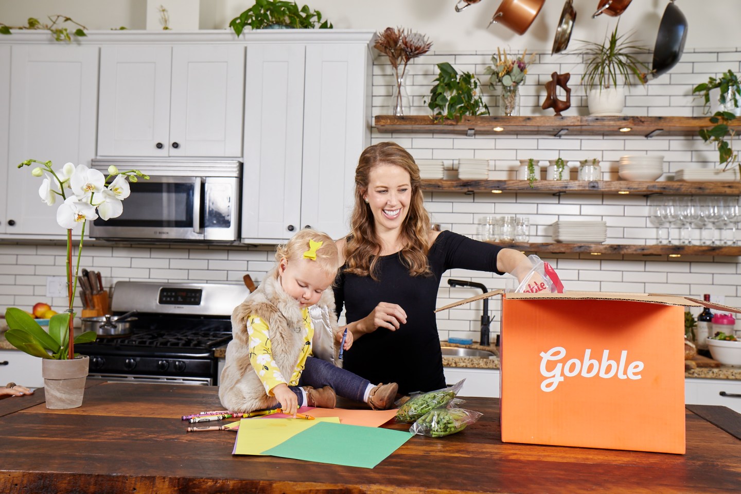 A woman opens a Gobble meal kit box while her young daughter sits on the counter and watches