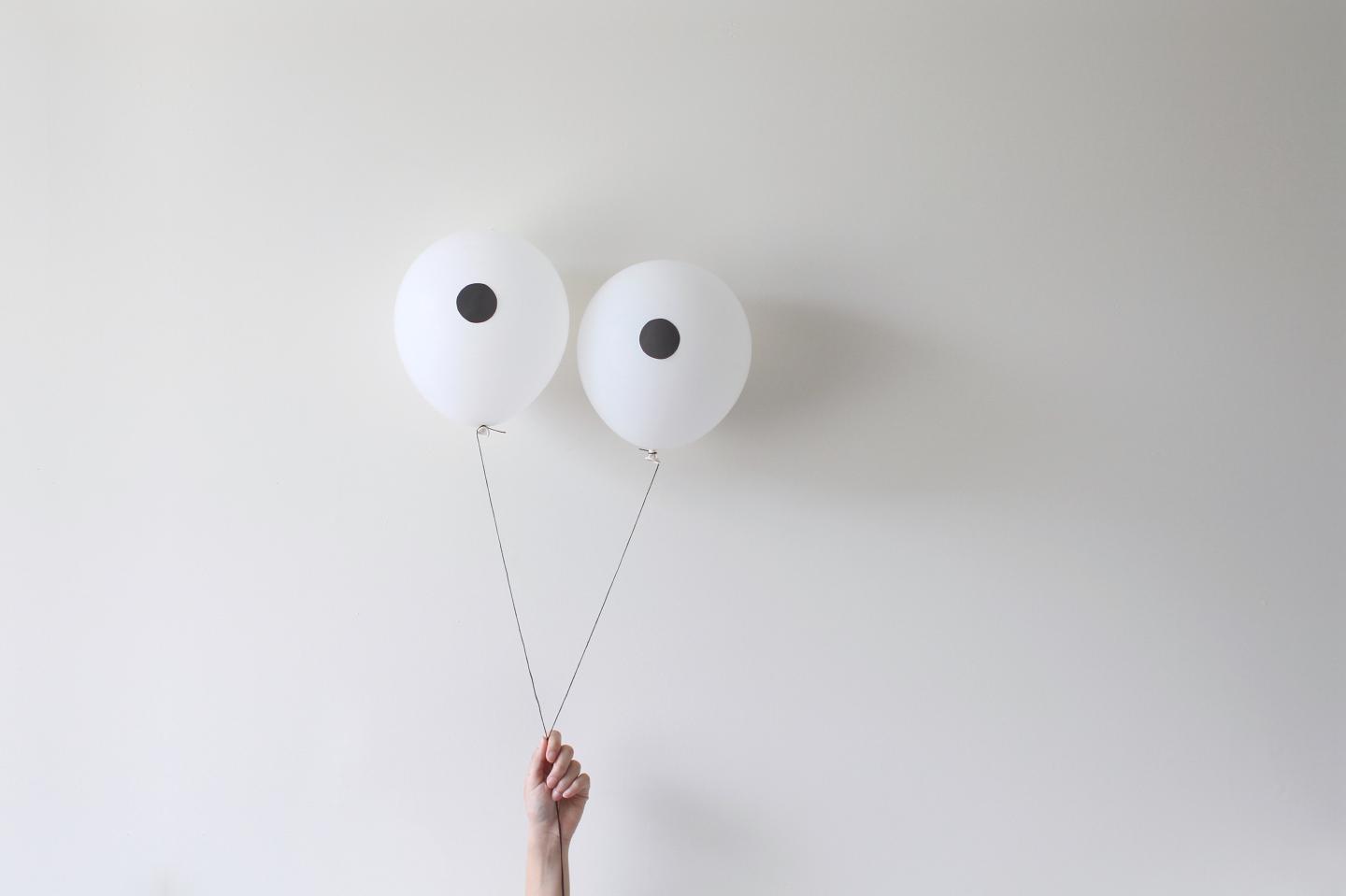 A hand holding a pair of balloons that look like eyes against a white wall