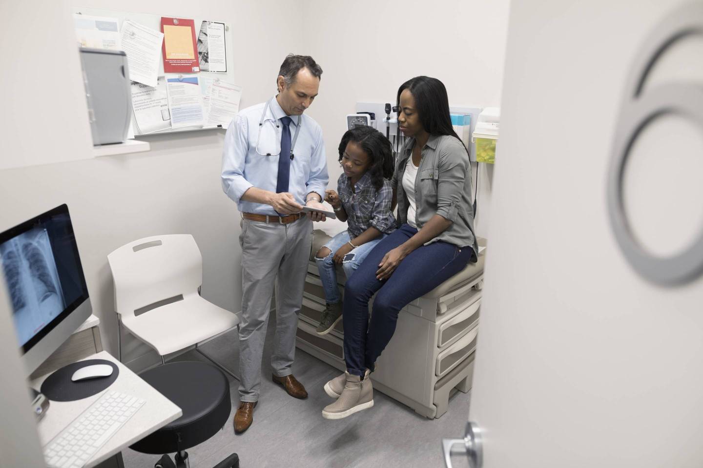 Male doctor with digital tablet talking to mother and daughter in clinic exam room