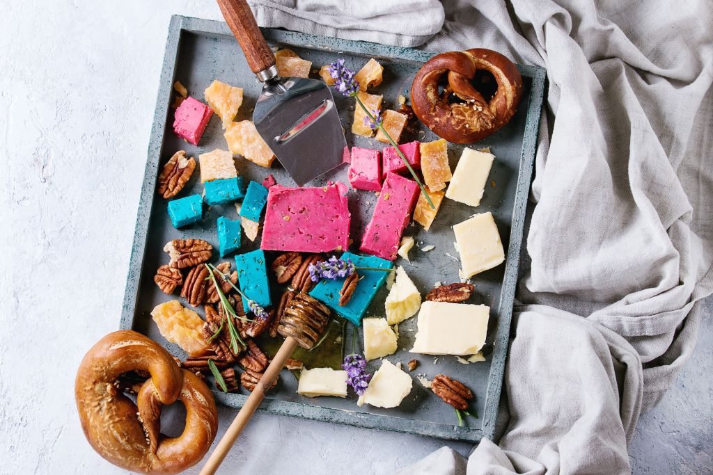 Variety of colorful holland cheese traditional soft, old, pink basil, blue lavender served with pecan nuts, honey, lavender flowers, pretzels bread on wood tray over gray texture background. Flat lay