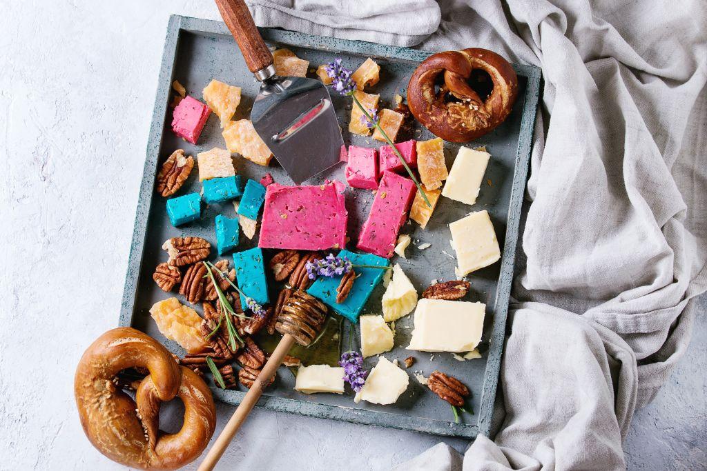 Variety of colorful holland cheese traditional soft, old, pink basil, blue lavender served with pecan nuts, honey, lavender flowers, pretzels bread on wood tray over gray texture background. Flat lay