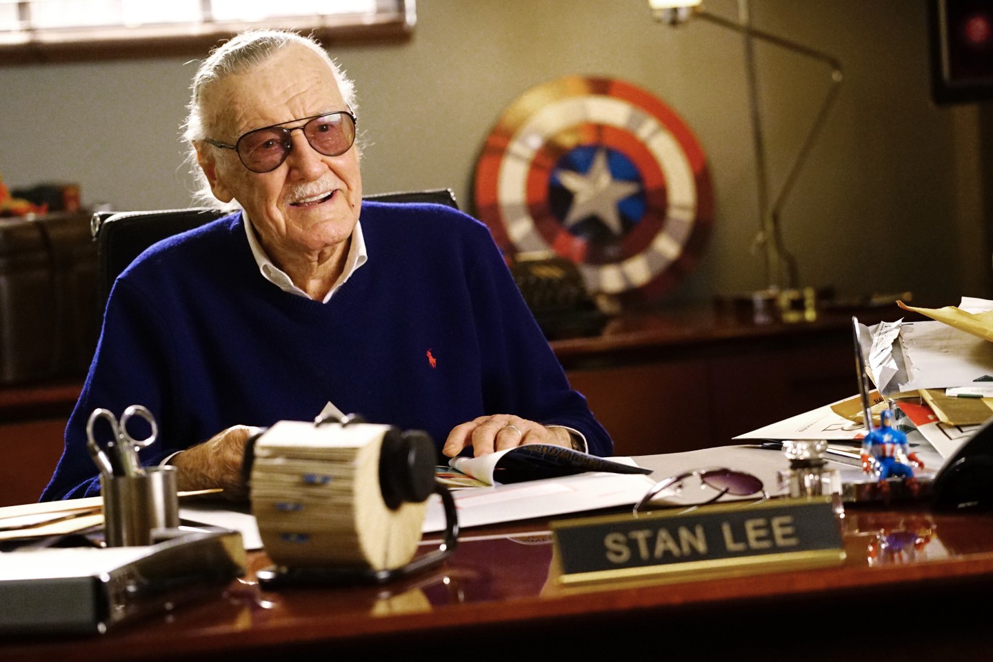 Stan Lee sits at a desk with a Captain America shield behind him on the set of ABC's FRESH OFF THE BOAT