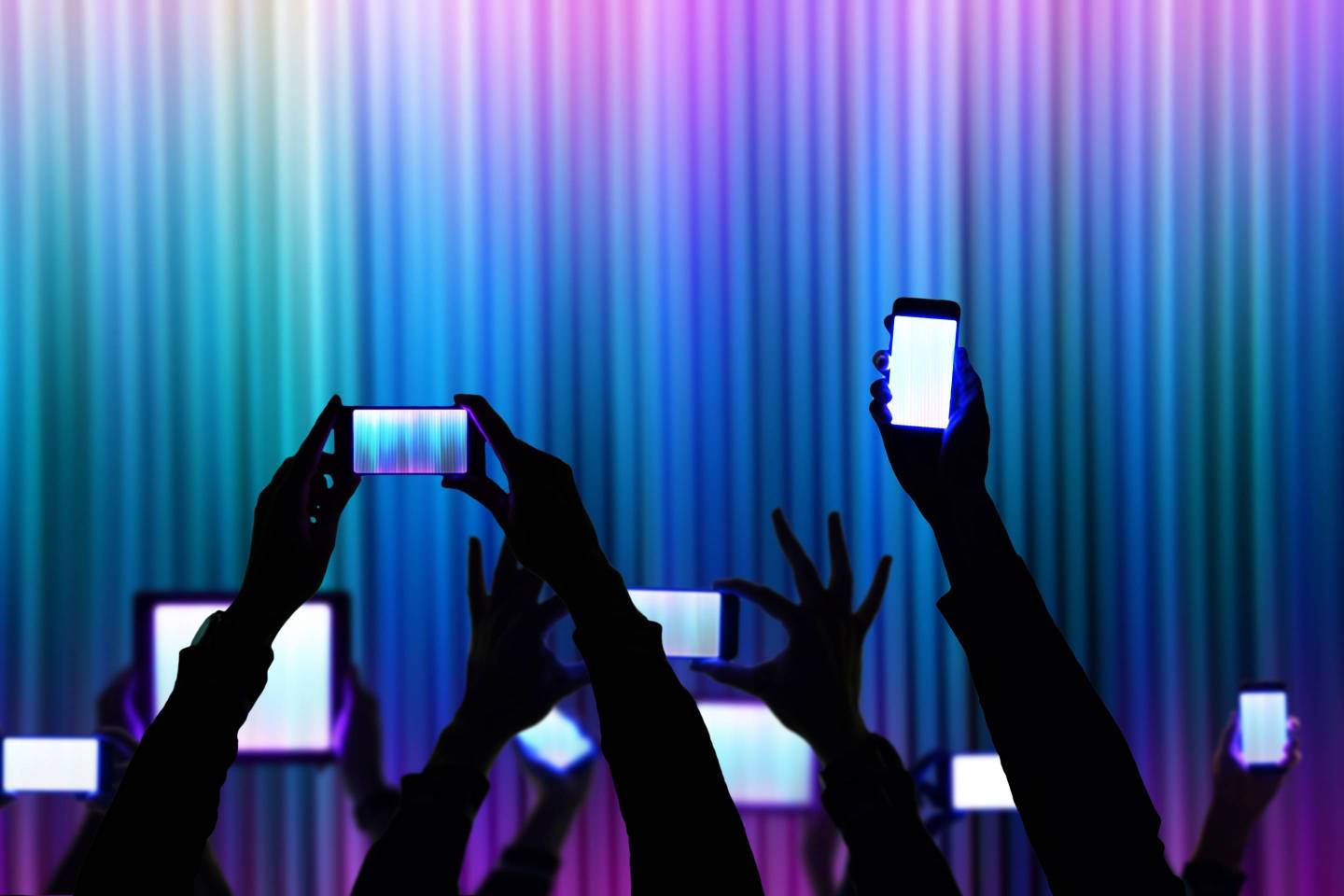 People raise his bright smartphone and tablet device during a night show celebration with dark silhouettes and colorful background.