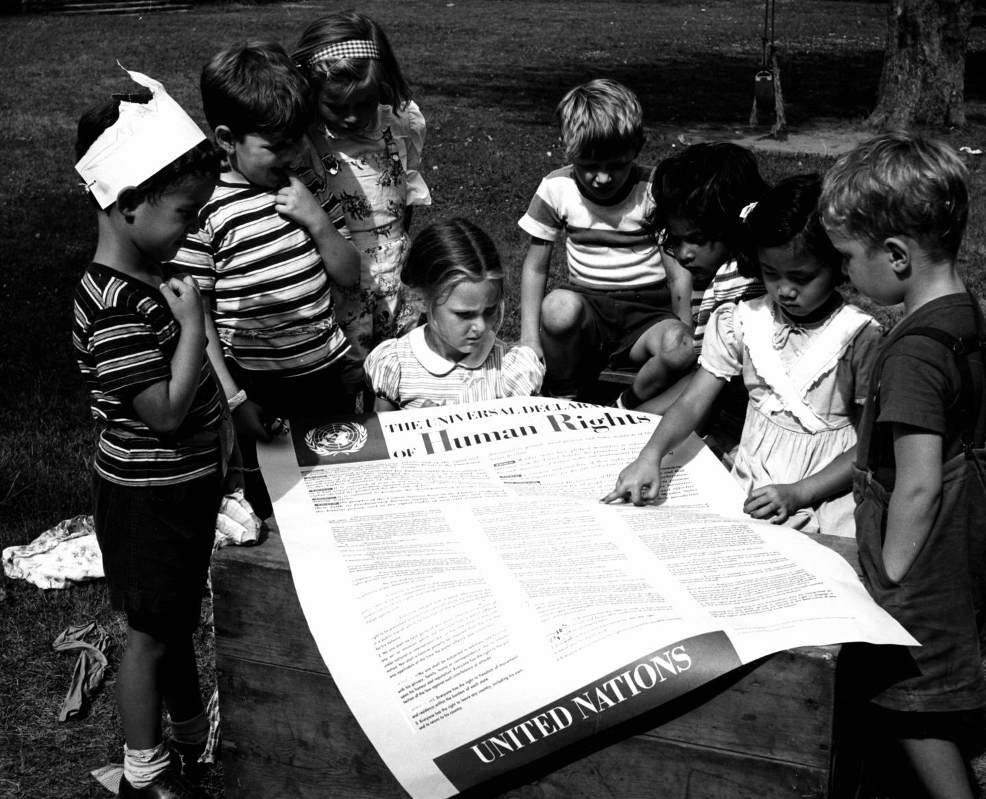 A group of school children sit outside, gathered around a large poster printed with the UN's Universal Declaration of Human Rights.