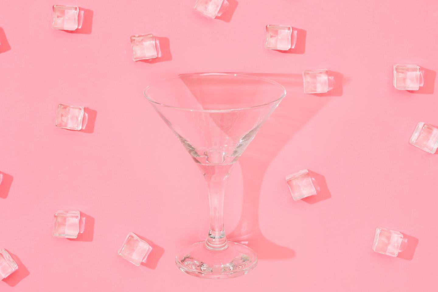 empty cocktail glass on a pink background surrounded by ice cubes.